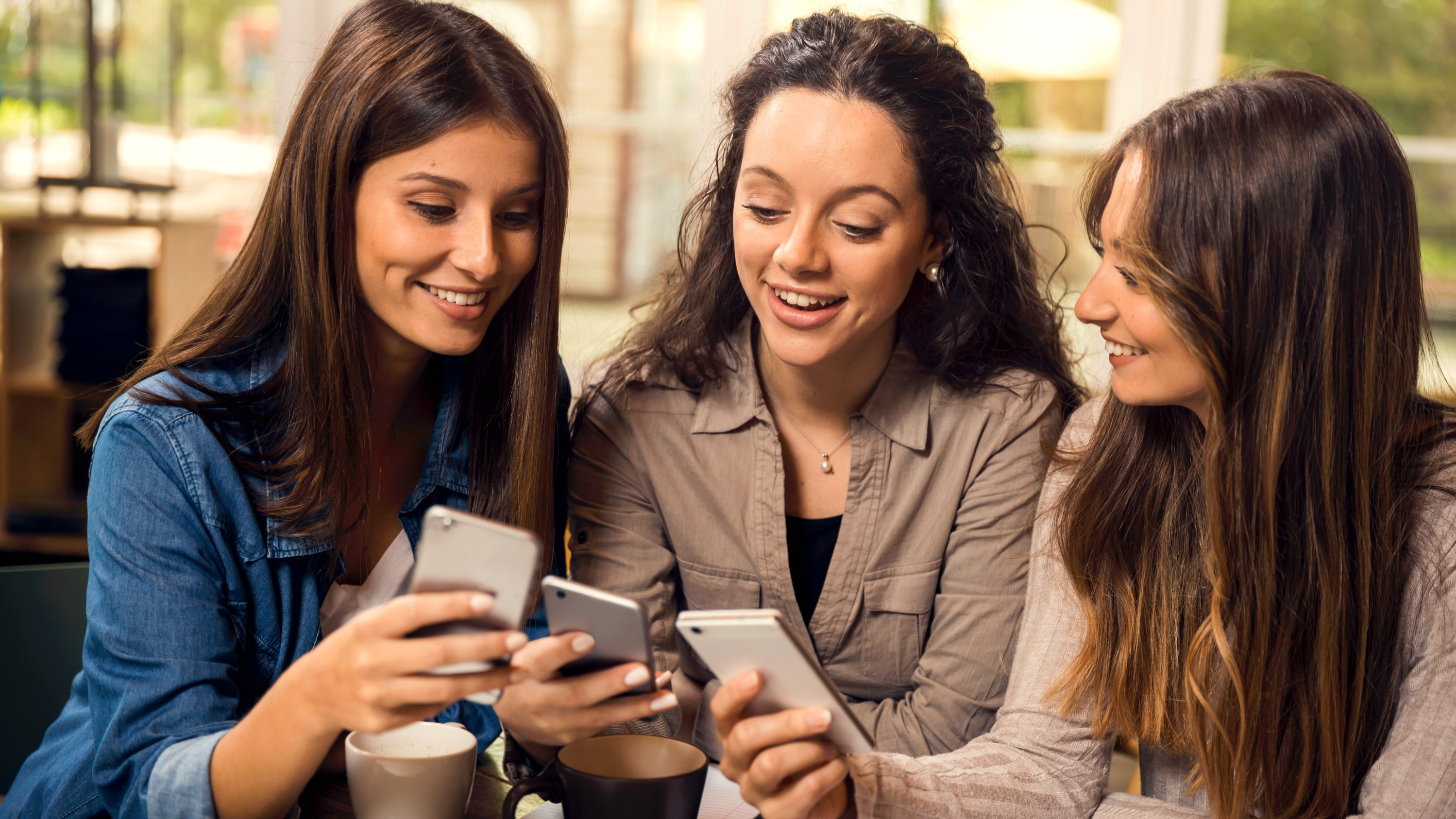 Unge kvinner på kafé ser på hverandres mobiltelefoner og smiler